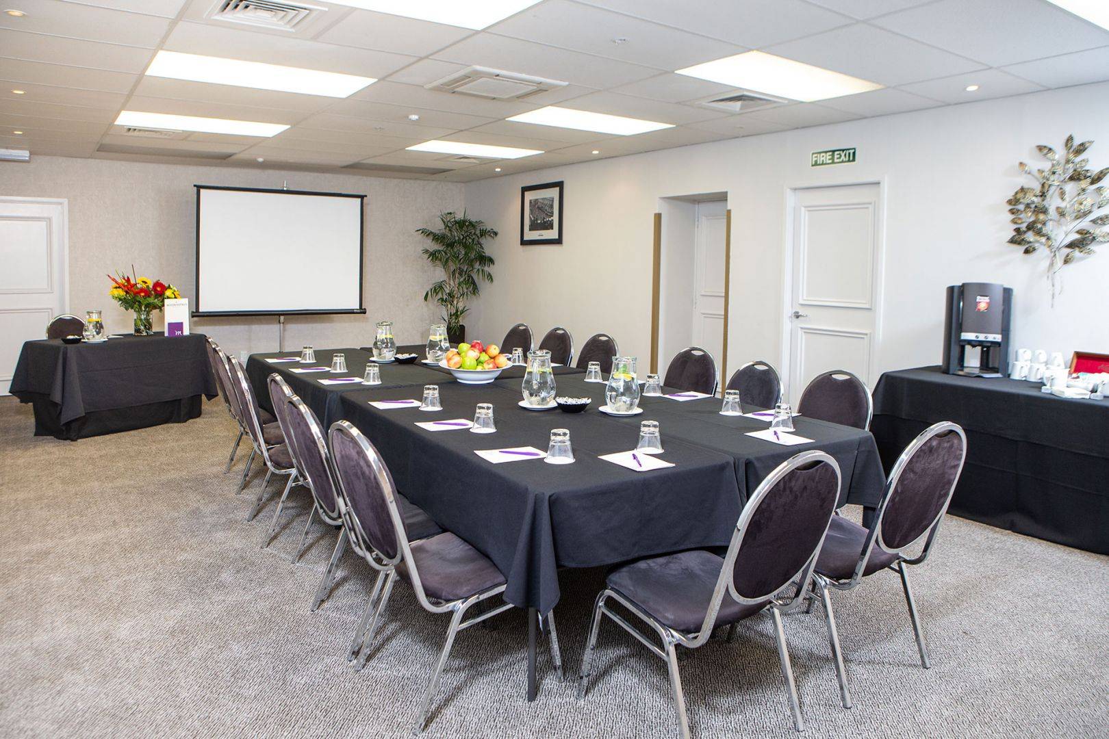 A windowless meeting room with a long table and chairs facing a projector screen on the wall.